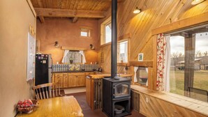 A rustic cabin interior with a wooden table, a black wood stove, a kitchen area with a refrigerator, and a window with curtains. - Off-Grid Cottage at Granny Flats (Spearfish)