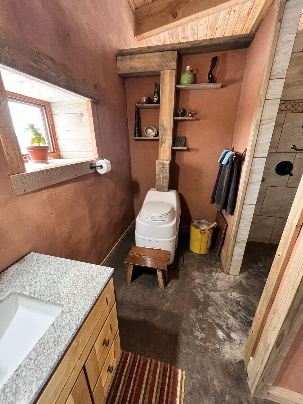 Une petite salle de bain comprenant des toilettes, un lavabo, des étagères en bois et une fenêtre agrémentée d’une plante.