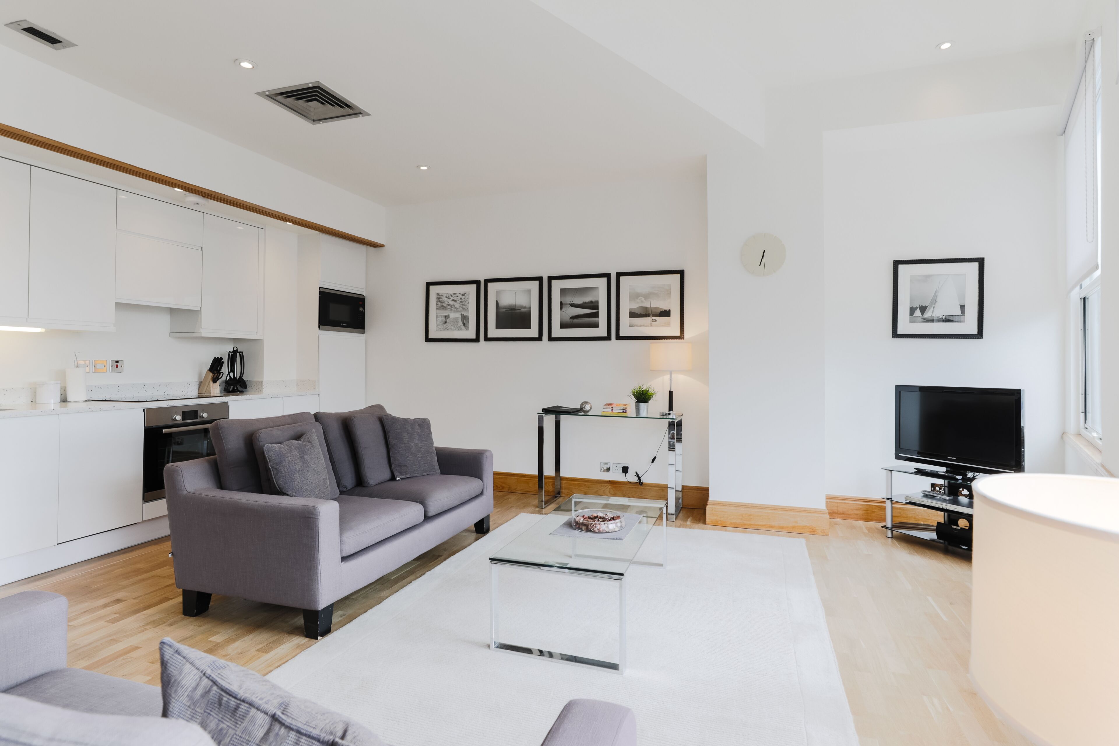 A modern living room with a grey sofa, a coffee table, and a TV. There is a kitchen area with white cabinets and a microwave.