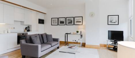 A modern living room with a grey sofa, a coffee table, and a TV. There is a kitchen area with white cabinets and a microwave.
