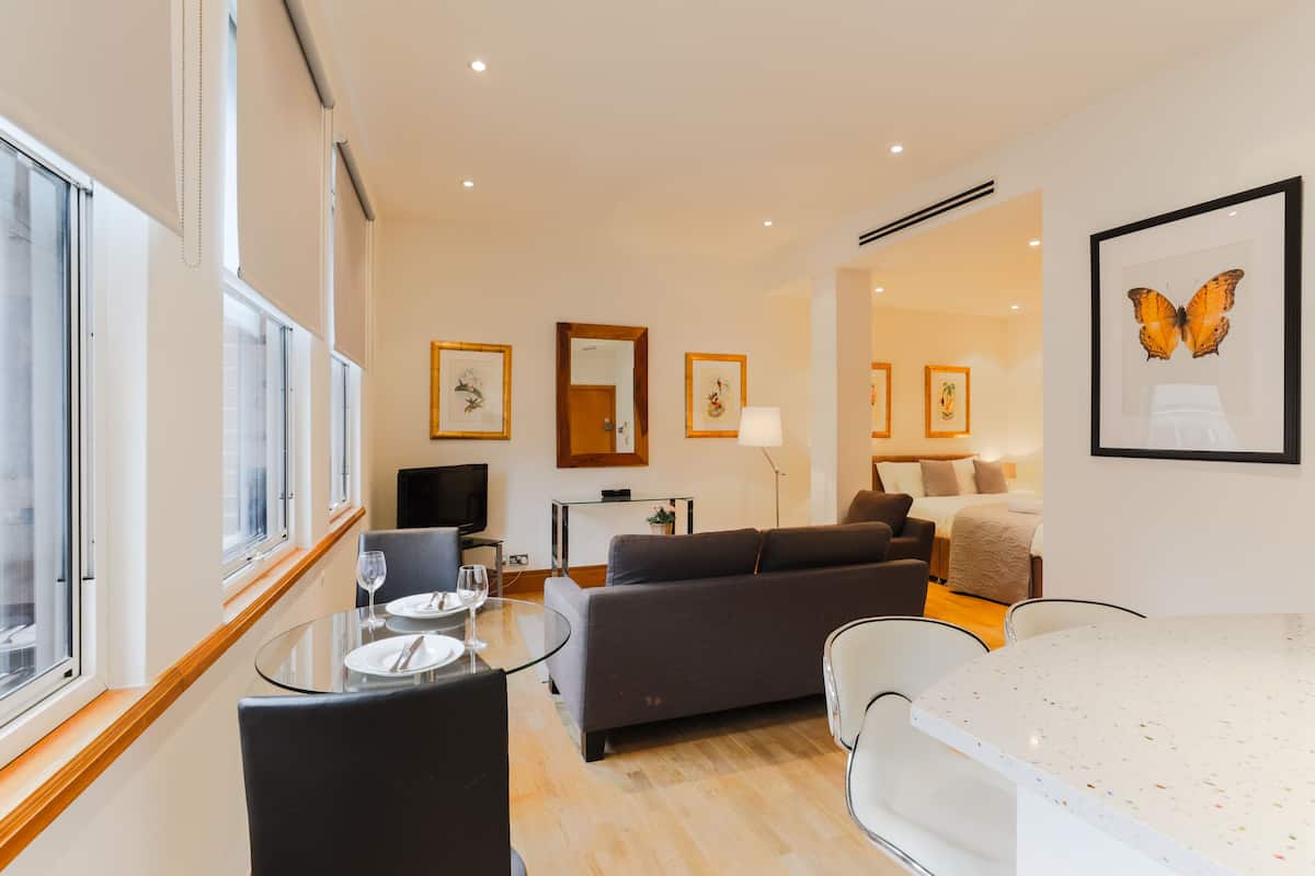 A modern living room with a sofa, a dining table, and a TV. There are framed pictures on the walls and a window with blinds.