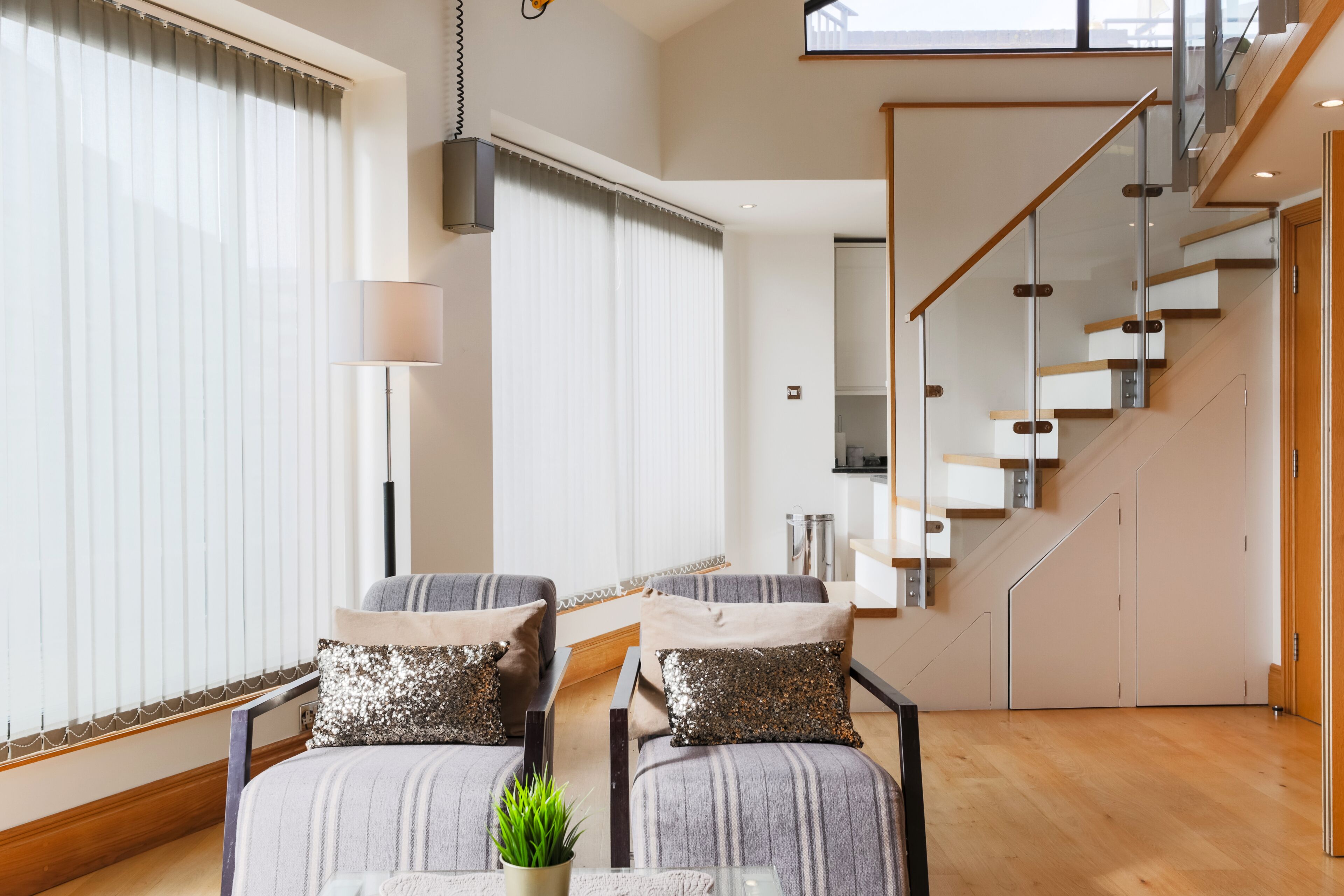 Ein modernes Wohnzimmer mit einer Sofagarnitur, einem Couchtisch und einer Topfpflanze. Eine Treppe mit Glastrennwand führt auf eine obere Ebene.