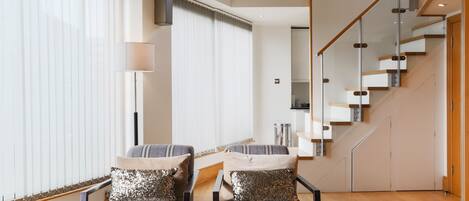 Ein modernes Wohnzimmer mit einer Sofagarnitur, einem Couchtisch und einer Topfpflanze. Eine Treppe mit Glastrennwand führt auf eine obere Ebene.