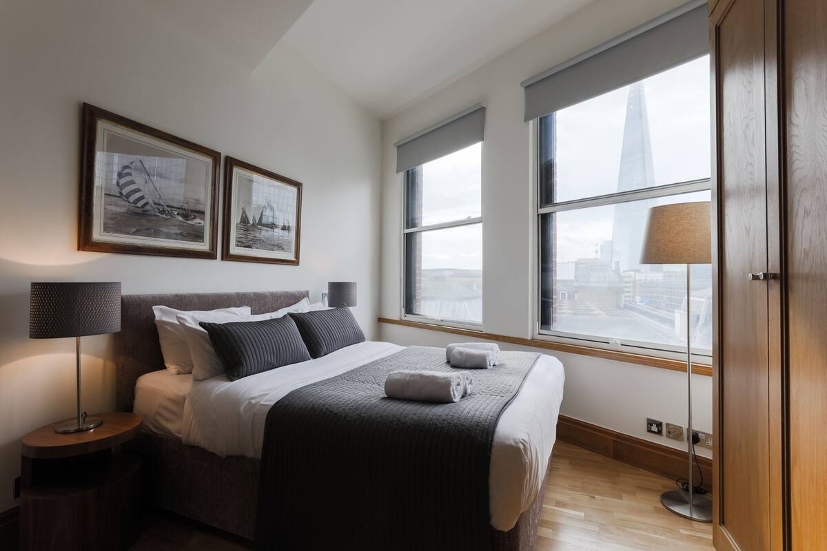 A modern hotel room with a large bed, two framed pictures on the wall, a window with a city view, and a bedside lamp.