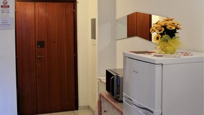 Habitación de hotel con puerta de madera, refrigerador blanco, microondas y una pequeña mesa con un jarrón de flores.