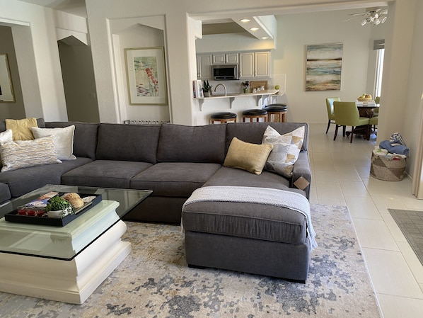 Una sala de estar moderna con un sofá seccional, un reposapiés y una mesa de centro de vidrio. Se observa una cocina al fondo con armarios blancos y una zona de comedor con sillas verdes.
