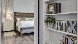 A bedroom with a wooden headboard, a white bed, a nightstand with a lamp, a white dresser, and a shelf with decorative items and books. - Water Well Wonder|Near Apple & Domain|Pet Friendly (Austin)