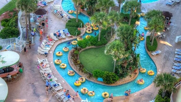 Un espace piscine doté de plusieurs toboggans, de chaises longues et d’une zone centrale couverte d’herbe, le tout entouré de palmiers.