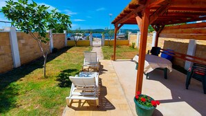 Ein überdachter Sitzbereich im Freien mit Tisch und Stühlen, ein kleiner Garten und Meerblick.
