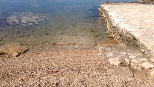 Ein Kiesstrand mit klarem Wasser, das zu einem Steg führt.