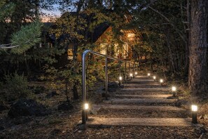 Un escalier en bois mène à un bâtiment entouré d’arbres, et des lampadaires illuminent le chemin.