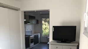 A modern kitchen with a stove, cabinets, and a window. There is a television on a stand and a bed with a striped blanket. - Studio Avec Piscine (Saint veran/Orgon)