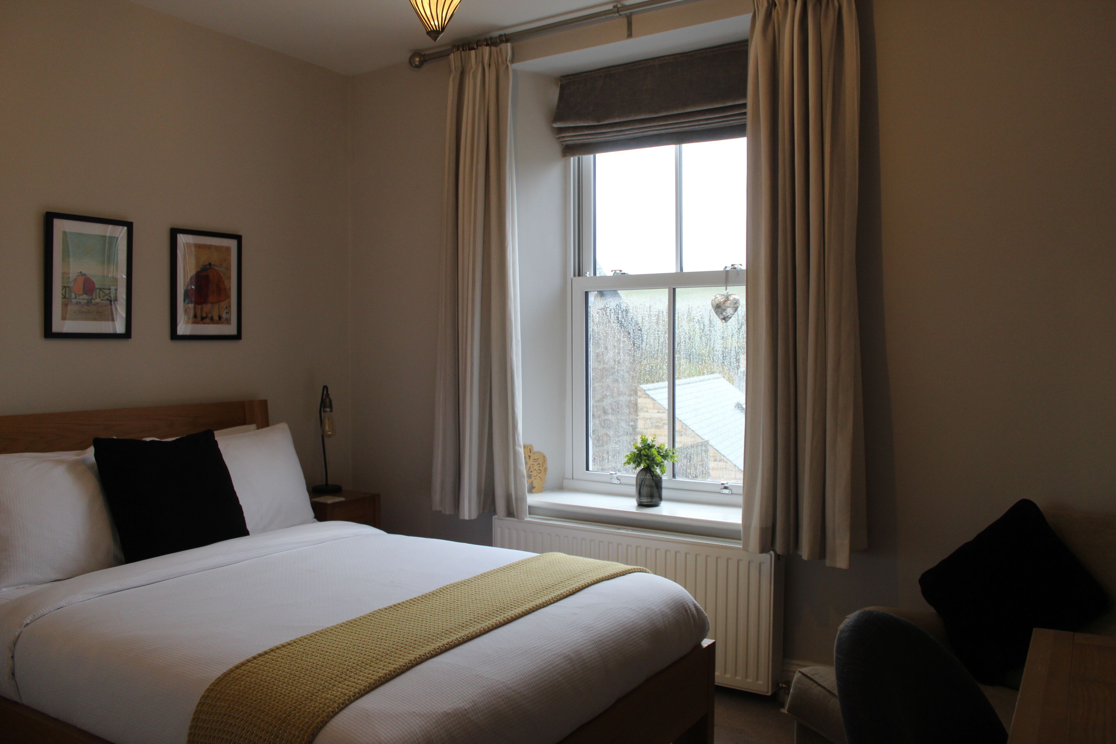Une chambre d’hôtel avec un grand lit, une fenêtre avec des rideaux, une table de chevet avec une lampe et deux tableaux encadrés au mur.