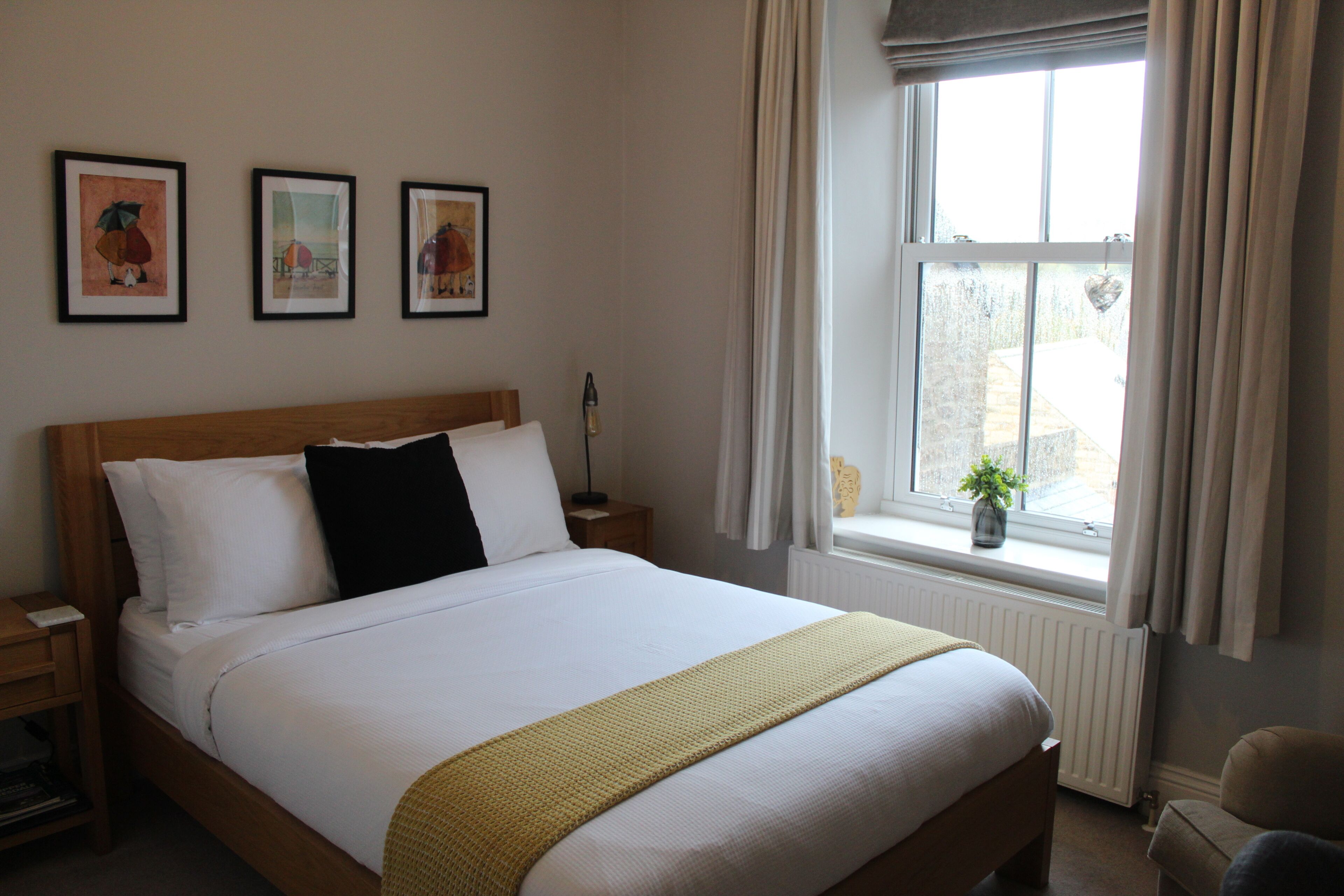 A neatly made bed with white linens and a yellow throw, a wooden headboard, and three framed artworks on the wall.