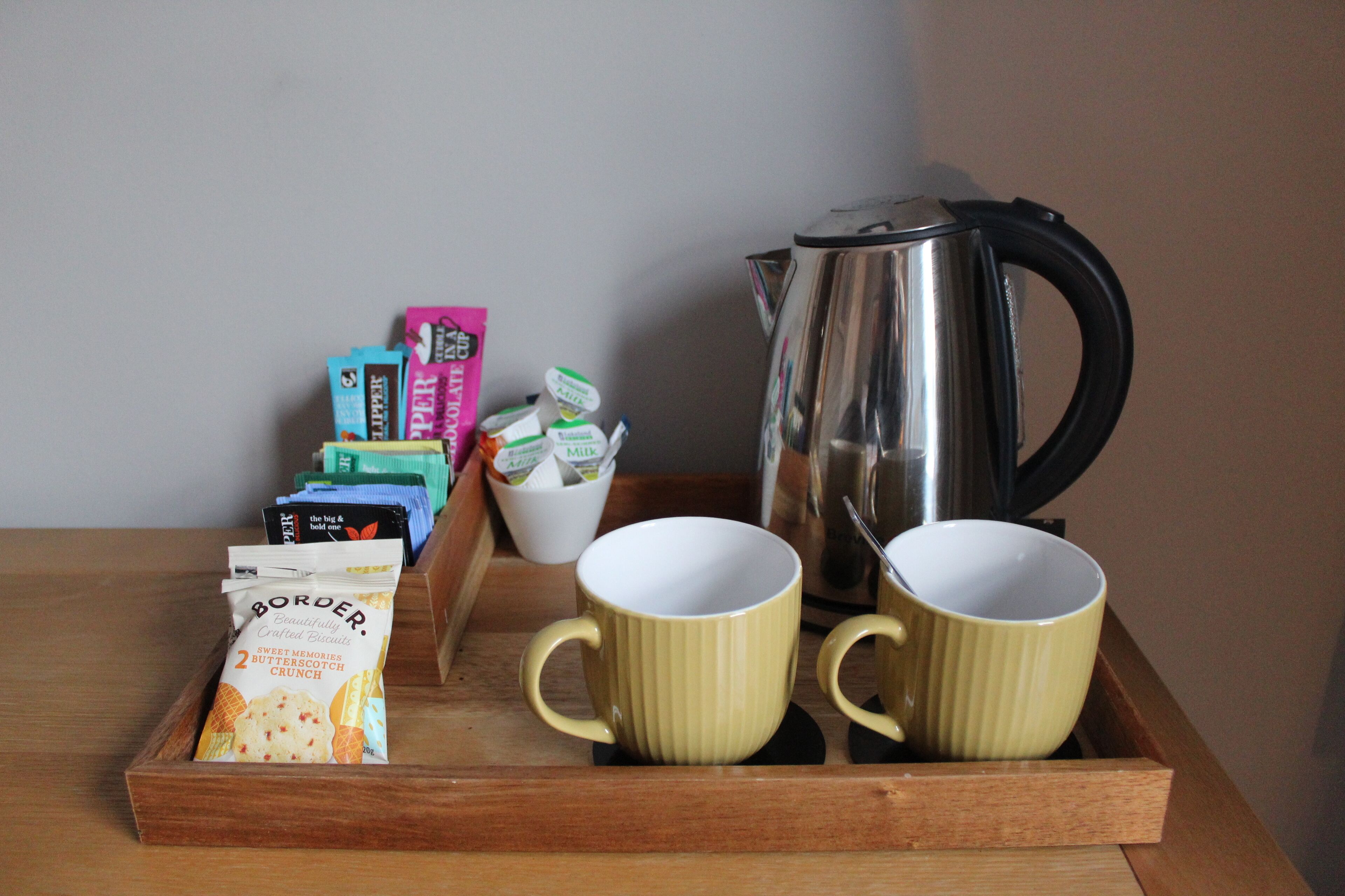 A tray with two yellow mugs, a silver electric kettle, and various packaged snacks.