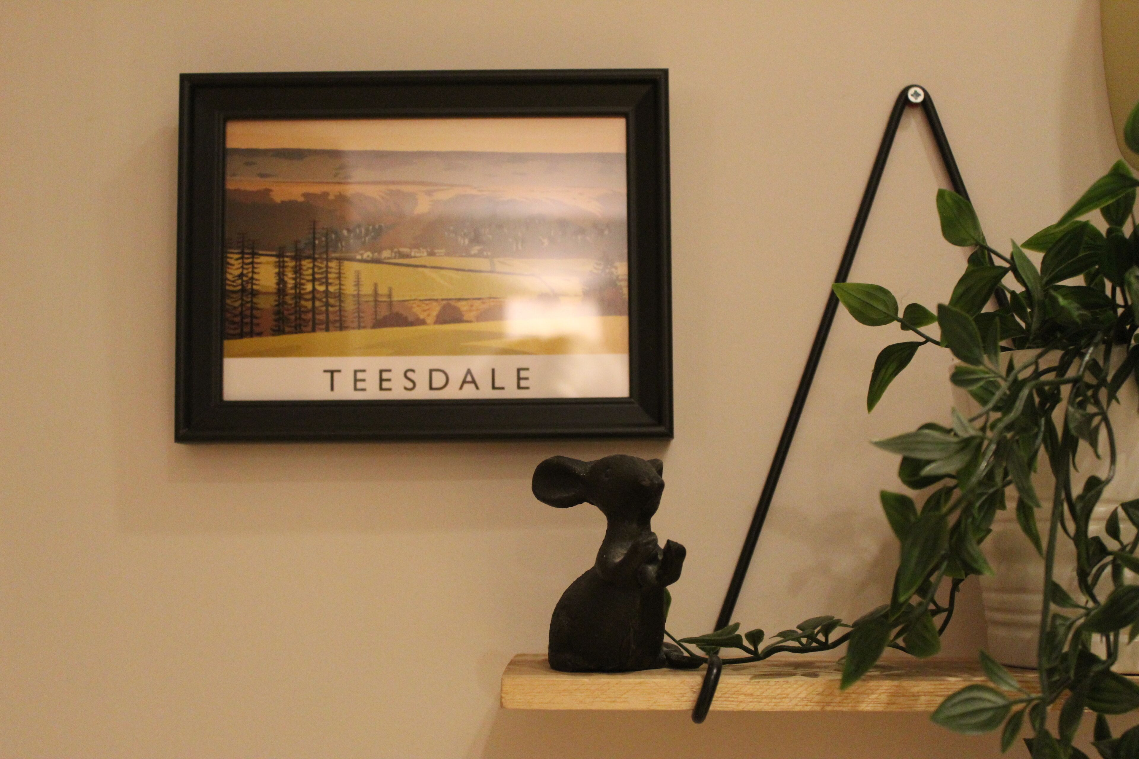 Un tableau encadré représentant Teesdale est accroché au mur, une petite figurine noire est posée sur une étagère, et une plante en pot se trouve à droite.