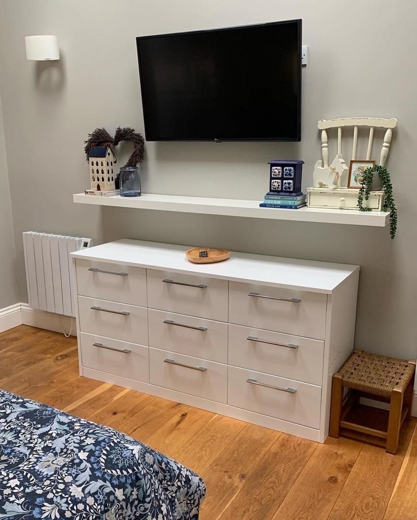Une pièce avec une commode blanche, un téléviseur fixé au mur au-dessus, et un petit tabouret en bois.