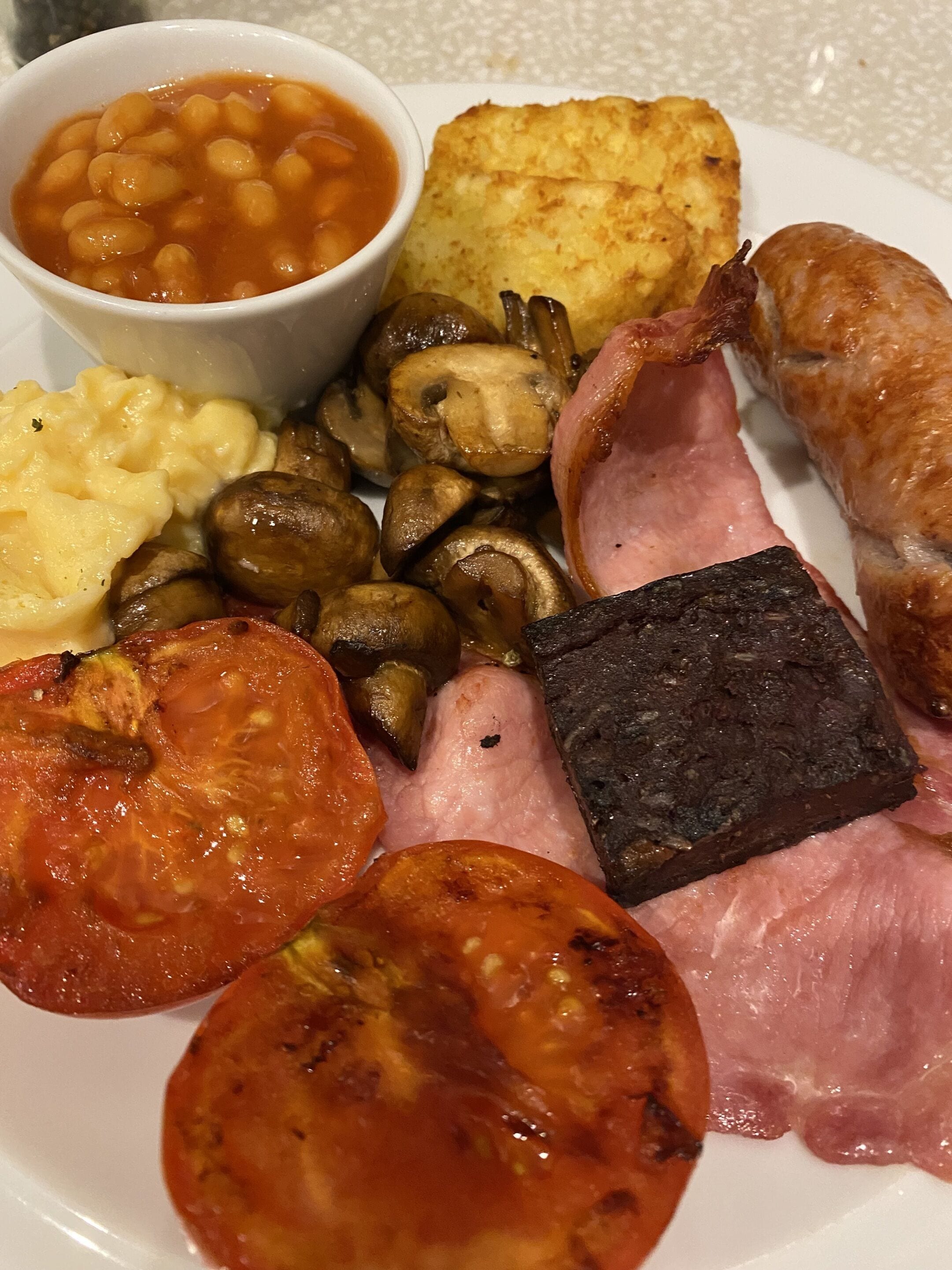 A plate of breakfast foods including beans, mushrooms, sausages, tomatoes, and ham.