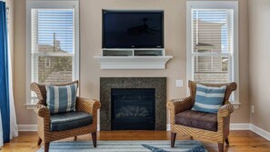 A living room with two wicker chairs, a fireplace, and a flat-screen TV mounted above. - Kure Sunrise, Private Townhome 1 Block From The Beach (Kure Beach)