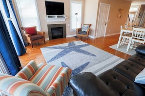 Una sala de estar con chimenea, televisor, alfombra con diseño de estrellas, sofás a rayas y de cuero, y un comedor con sillas blancas.