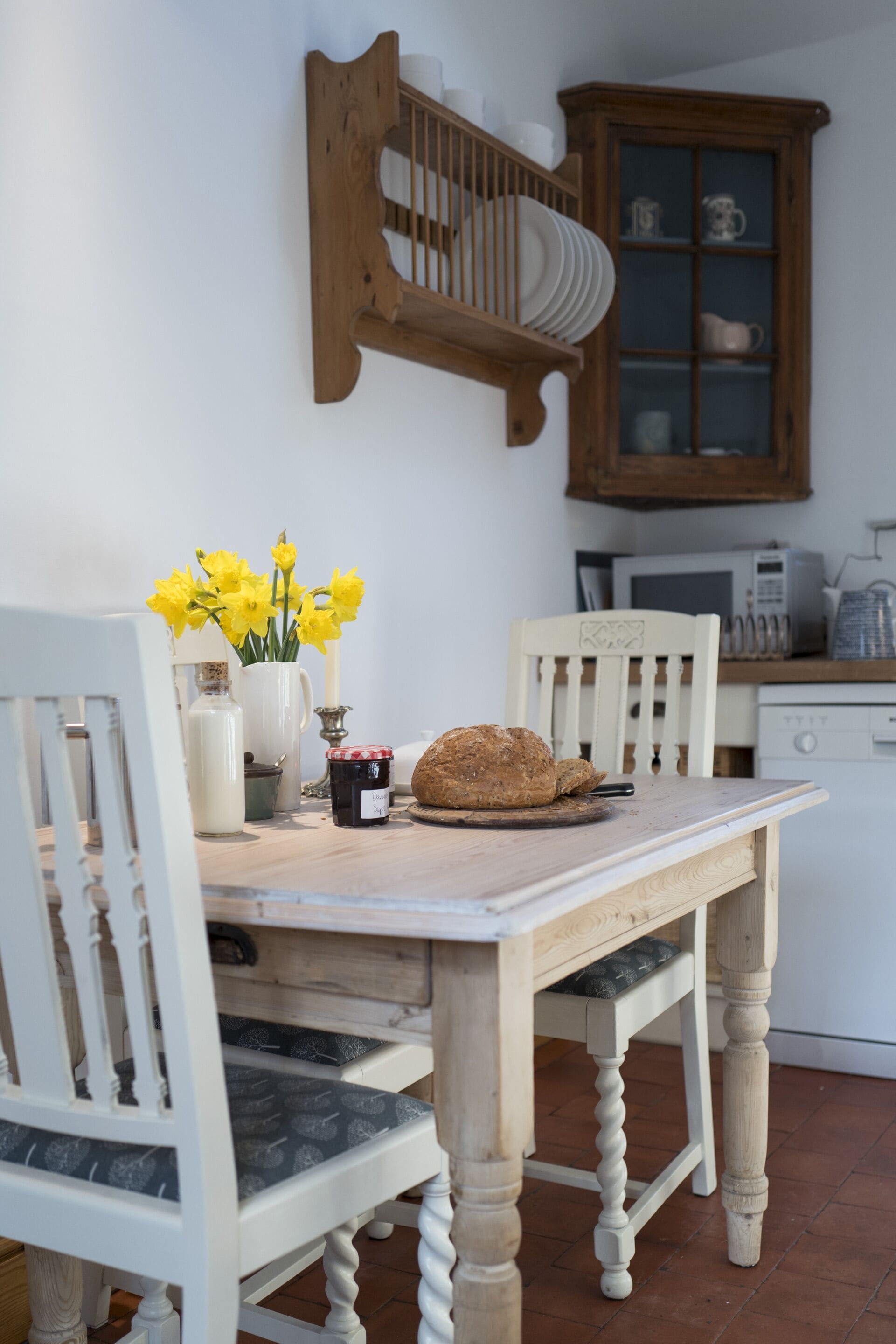 A dining table with chairs, a vase of yellow flowers, a loaf of bread, and a jar of jam.