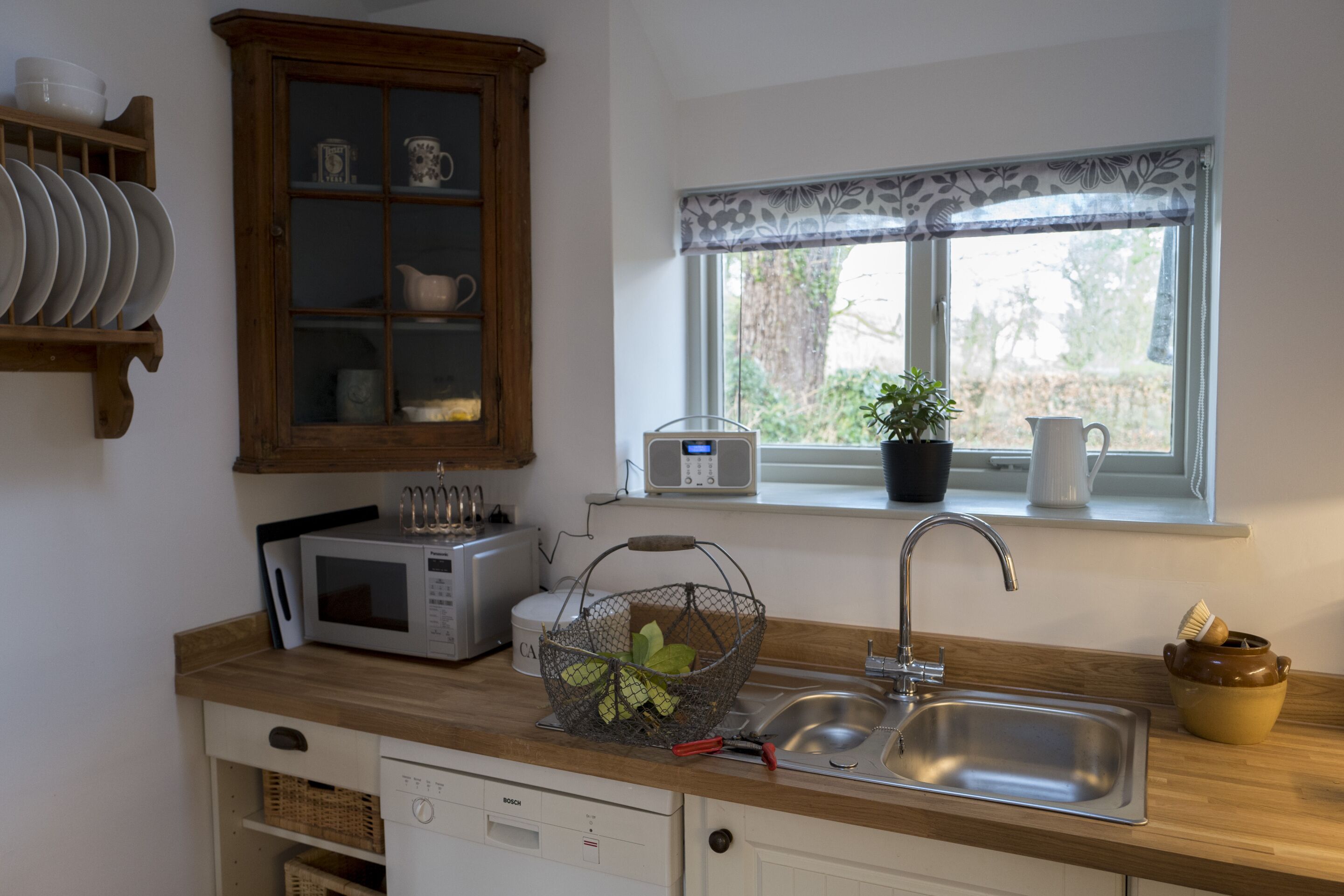 A kitchen with a wooden cabinet, a microwave, a sink, and a window with a plant.