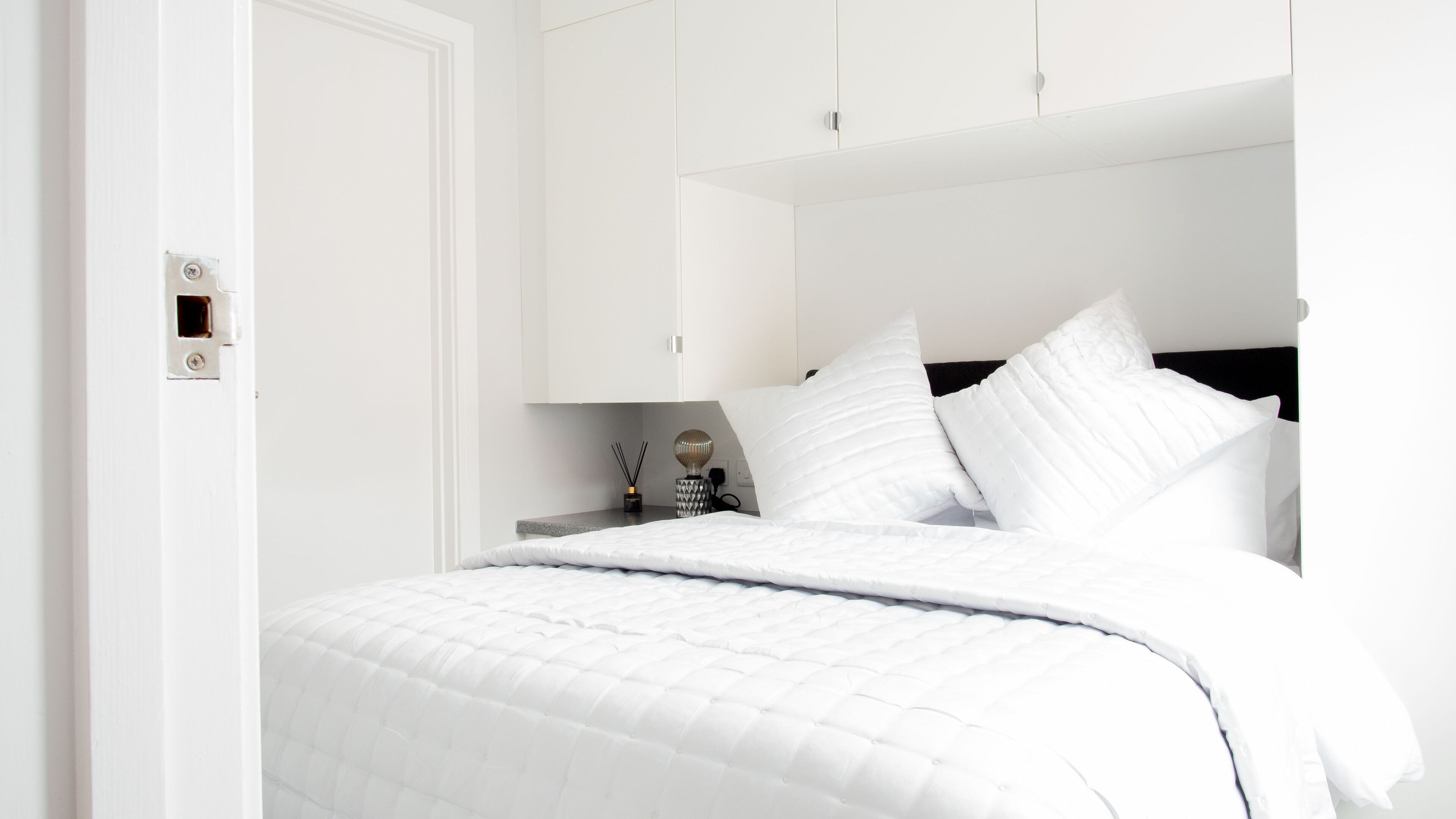 Un lit bien fait, avec une literie blanche, une table de chevet avec une lampe et un meuble fixé au mur.