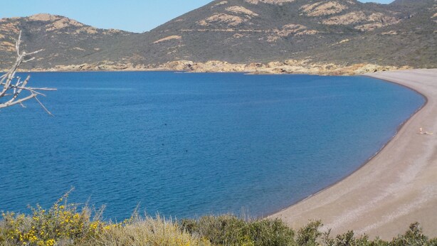 Un paisaje costero con mar azul cristalino, playa de arena y terreno montañoso al fondo.