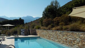 Una piscina con paredes de piedra, rodeada de sillones de baño y sombrillas, con montañas al fondo.