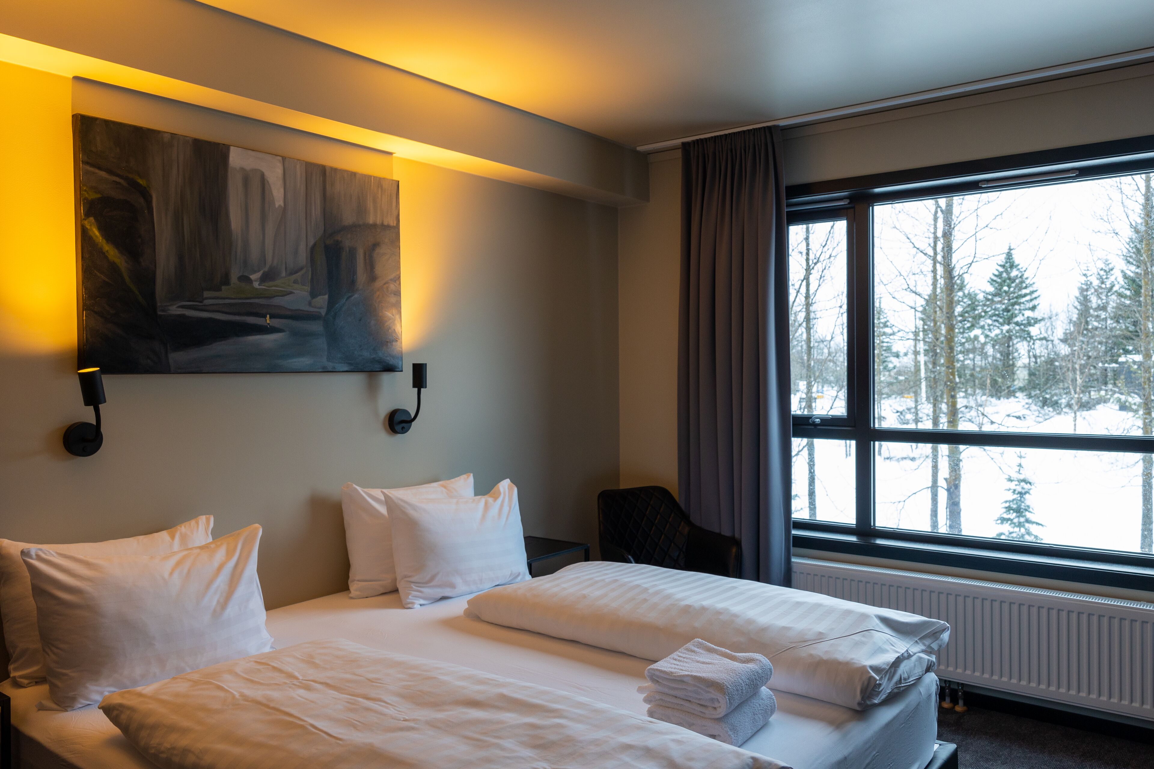 Une chambre d’hôtel avec un lit, des oreillers, un tableau, une chaise et une fenêtre donnant sur des arbres.