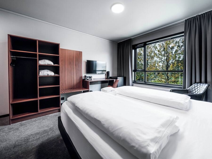 Habitación de hotel con una cama grande, un armario, un escritorio con computadora, televisor y ventana con vista a árboles.