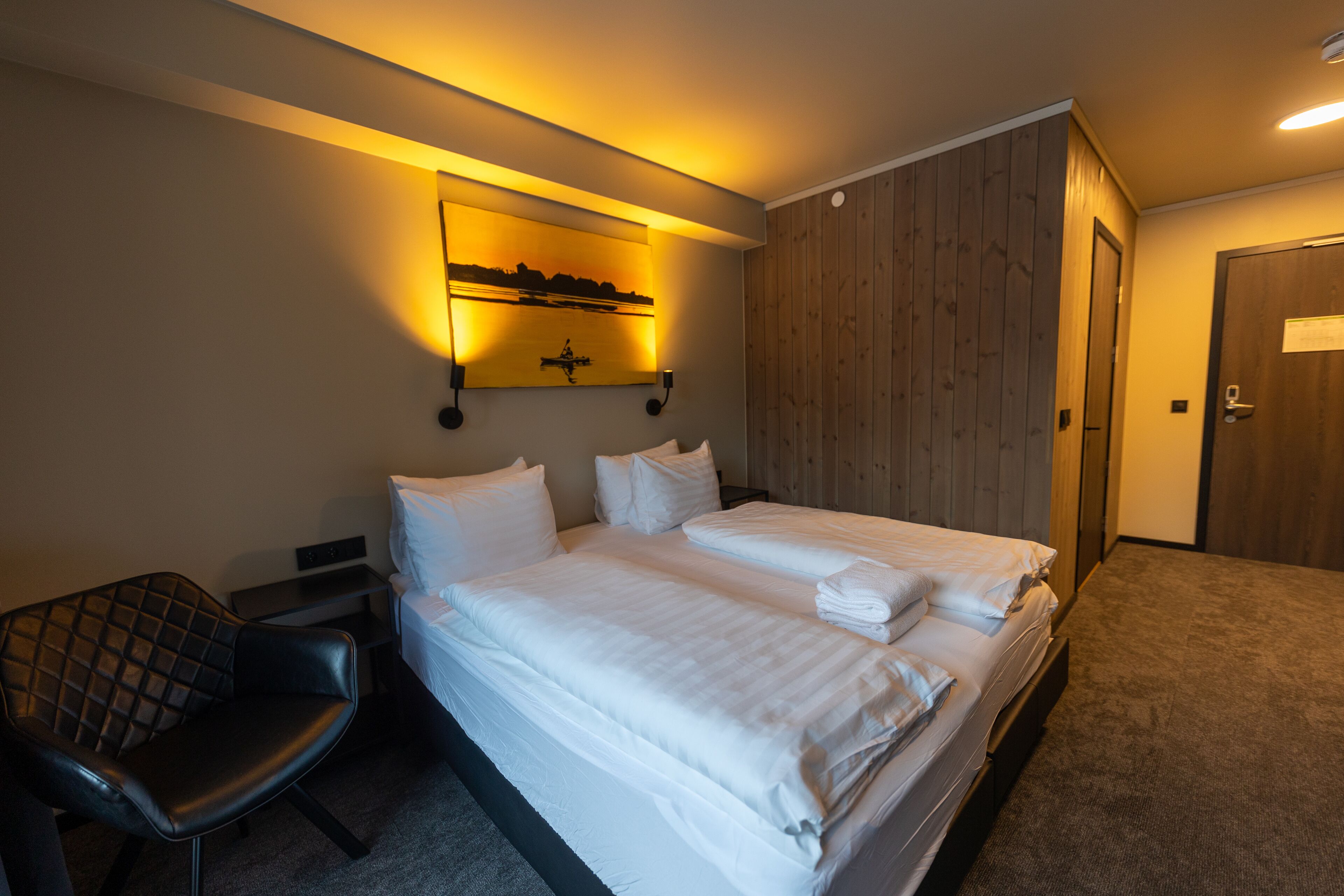 Habitación de hotel con dos camas, una silla, una obra de arte colgada en la pared y un panel de pared de madera.