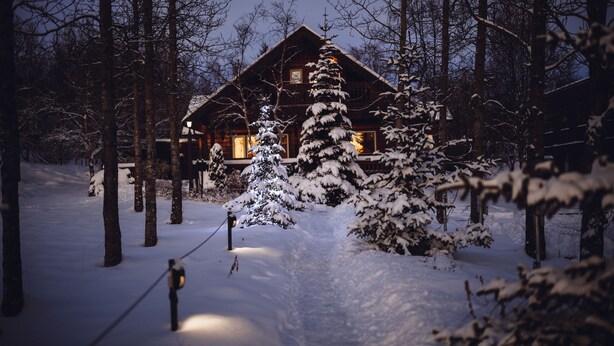 Un sentier enneigé menant à un chalet en bois dont une fenêtre est illuminée, le tout entouré d’arbres chargés de neige.