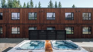 Un bâtiment moderne doté d’une façade en bois et de nombreuses fenêtres. Devant, on trouve une terrasse en bois avec deux bains à remous.