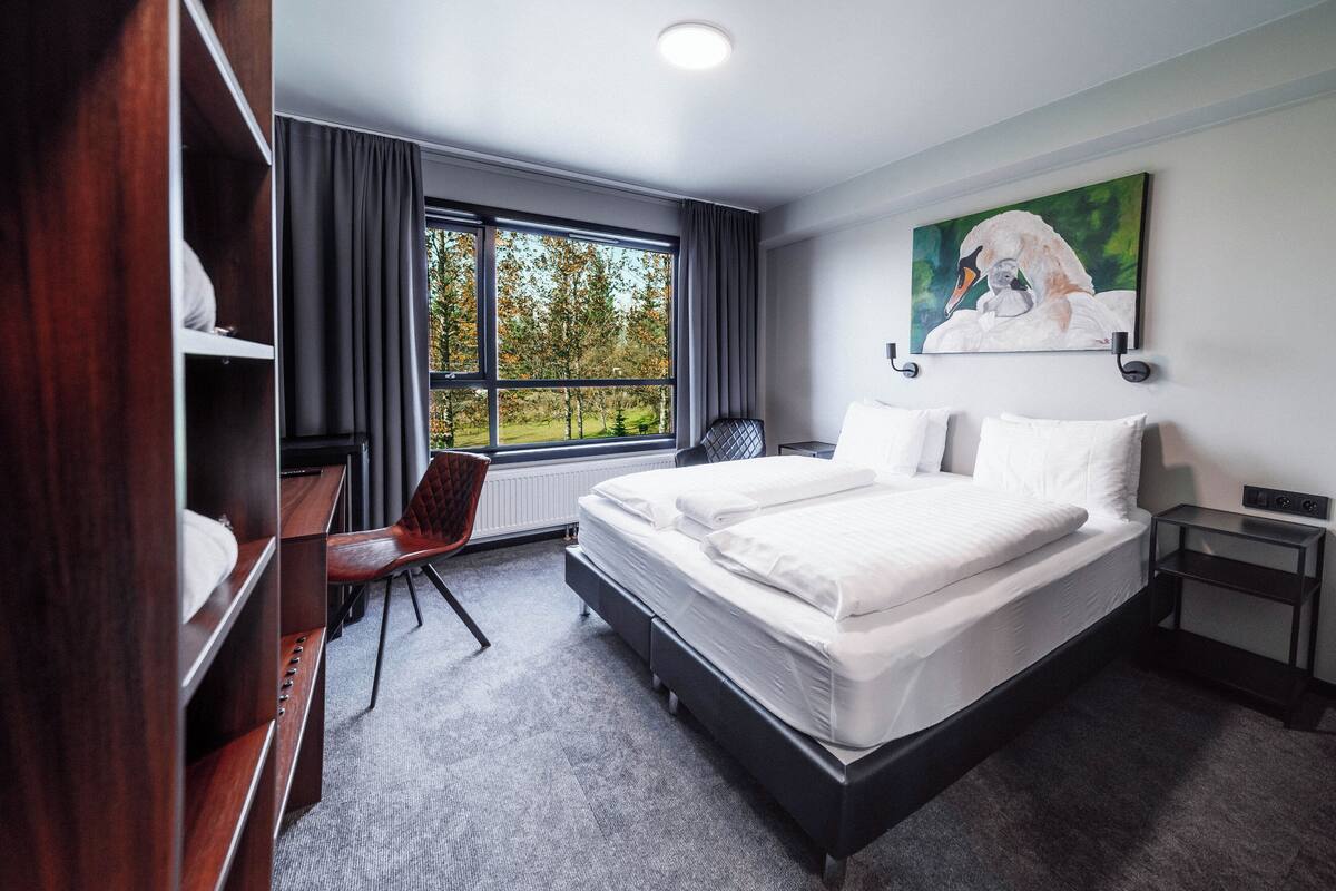 Una habitación de hotel con cama, escritorio, silla, ventana con cortinas y un cuadro en la pared.