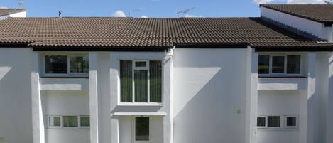 A two-story residential building with a tiled roof, white exterior walls, and a small front garden with potted plants.