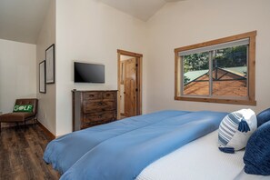 Un dormitorio con cama, televisor en la pared, cómoda de madera, silla con almohada de golf y ventana con vista a una casa.