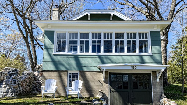Une maison à deux étages avec une façade en pierre, portant le numéro 180, dotée d’un porche avec deux fauteuils Adirondack blancs et d’un muret en pierres.