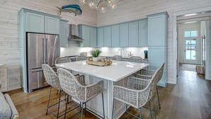 A modern kitchen with light blue cabinets, a stainless steel refrigerator, wicker chairs, and a chandelier.