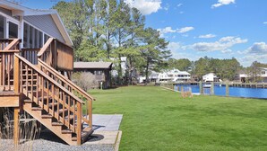 A waterfront property with a wooden deck, a grassy lawn, and a clear view of the water. - Our Happy Place | Canal Front w/ Private Dock, Huge Screened Porch + Pet Friendly Escape (Chincoteague Island)