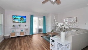 A living room with a wooden floor, a white sofa, a TV mounted on the wall, a ceiling fan, and a sliding glass door leading to a balcony. - Our Happy Place | Canal Front w/ Private Dock, Huge Screened Porch + Pet Friendly Escape (Chincoteague Island)