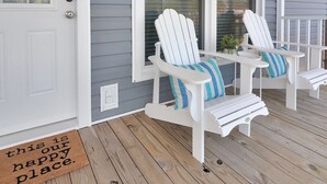 A porch with two Adirondack chairs and a small table, a doormat with a message, and a white door. - Our Happy Place | Canal Front w/ Private Dock, Huge Screened Porch + Pet Friendly Escape (Chincoteague Island)