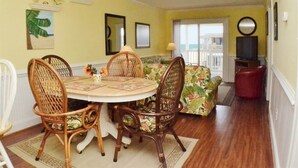 A dining area with a round table and wicker chairs, a ceiling fan, and a view of the balcony. - Oceanfront Condo on the Outer Banks (Kill Devil Hills)
