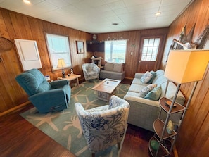A cozy living room with wooden walls, a blue armchair, a patterned armchair, a grey sofa, a coffee table, and a floor lamp. - Charming Oceanfront retreat (Kill Devil Hills)