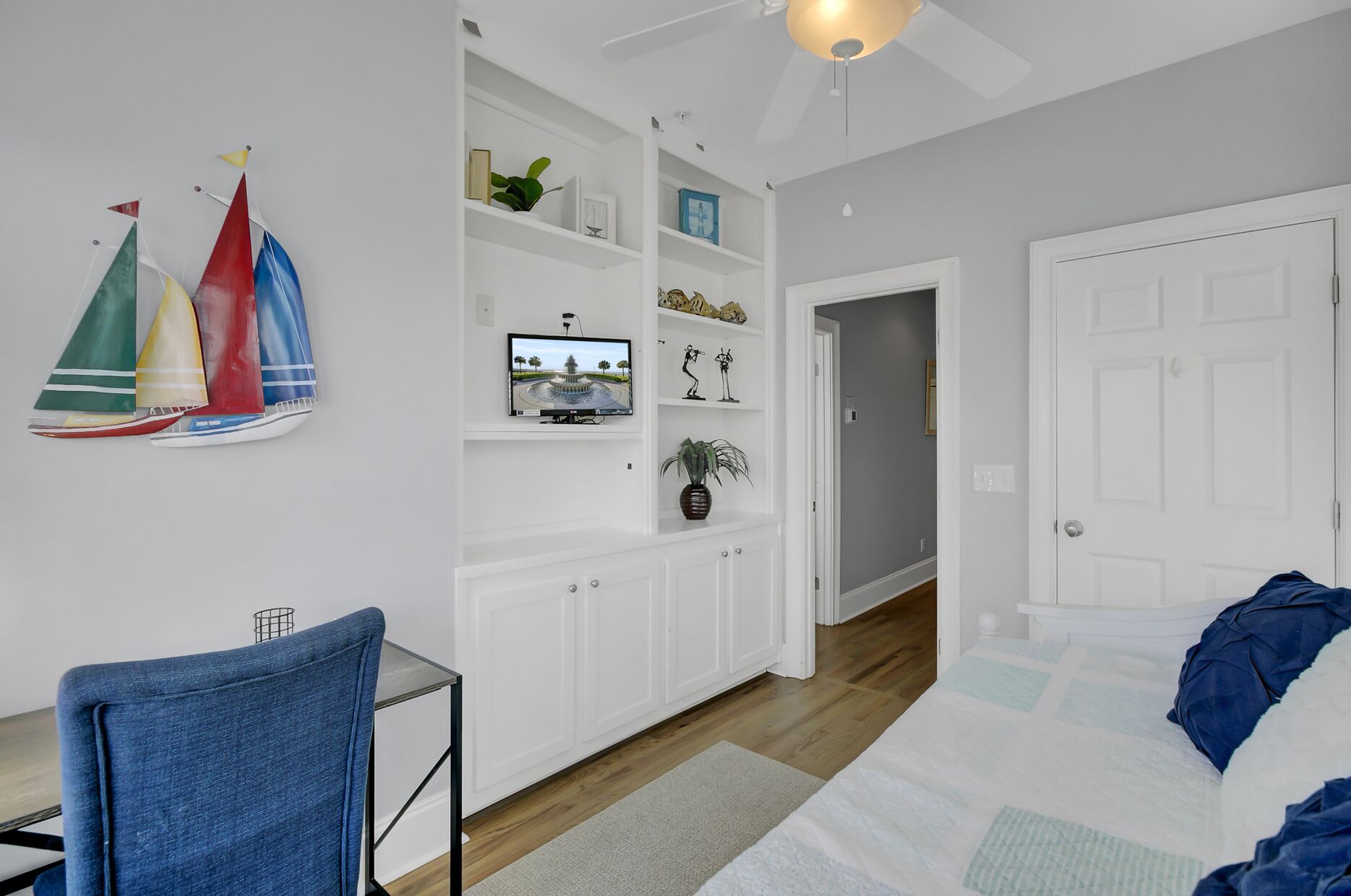 A bedroom with a bed, a desk, and a chair. There is a white door and a hallway leading to another room.