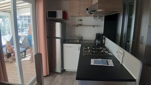 A compact kitchen with a microwave, refrigerator, and stove. There is a sliding door leading to an outdoor area with a deck. - Loue Mobilhome (Sérignan)