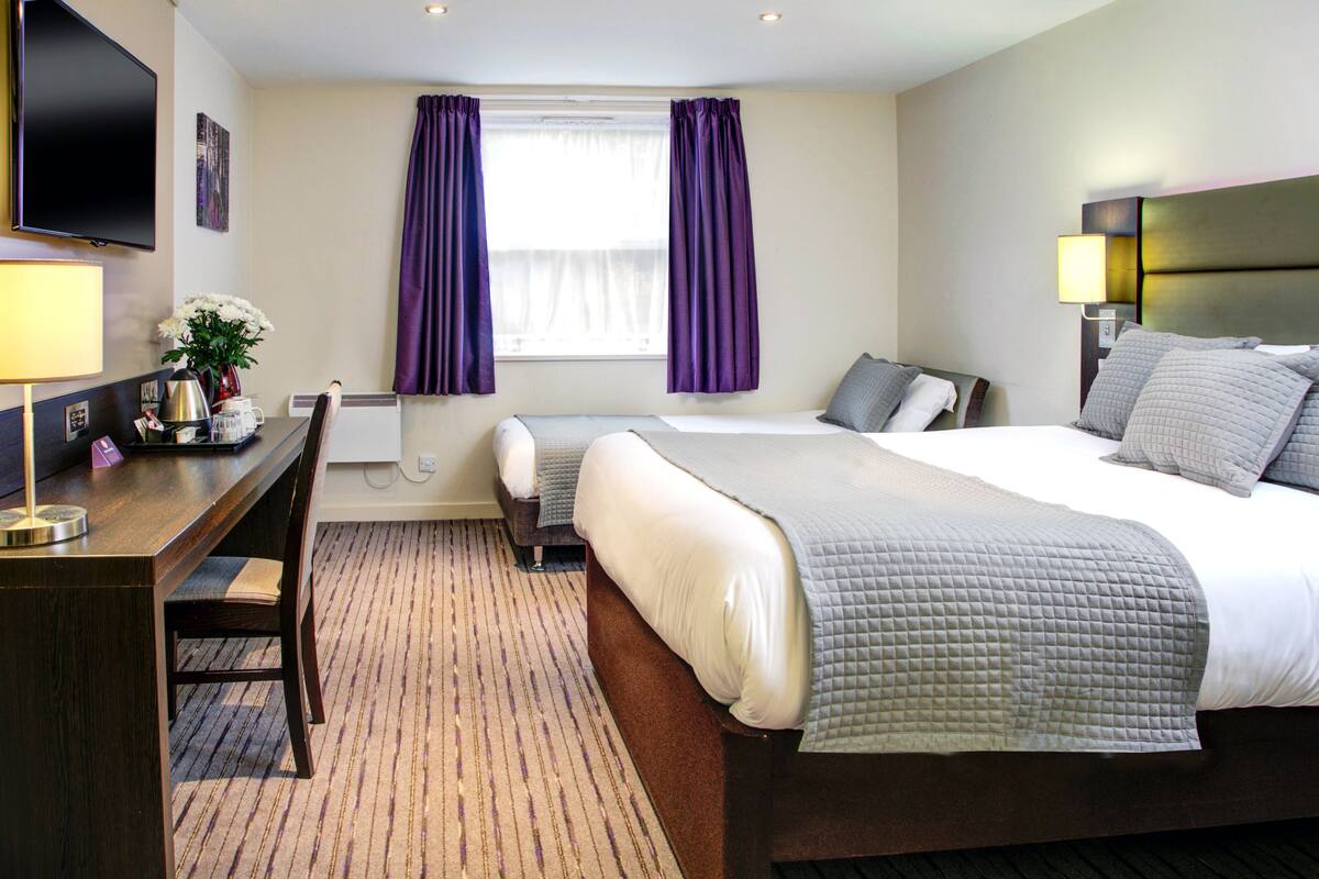 A hotel room with a large bed, a smaller bed, a desk with a chair, a TV, and a window with purple curtains.