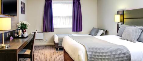 Habitación de hotel con una cama grande, una cama más pequeña, un escritorio con silla, televisión y ventana con cortinas moradas.
