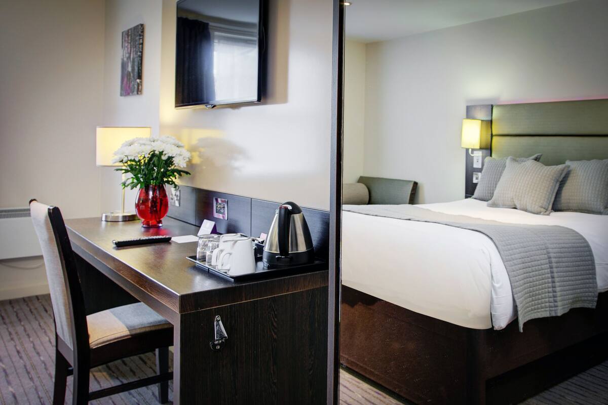 A hotel room with a bed, a desk with a vase of flowers, a television, and a lamp.