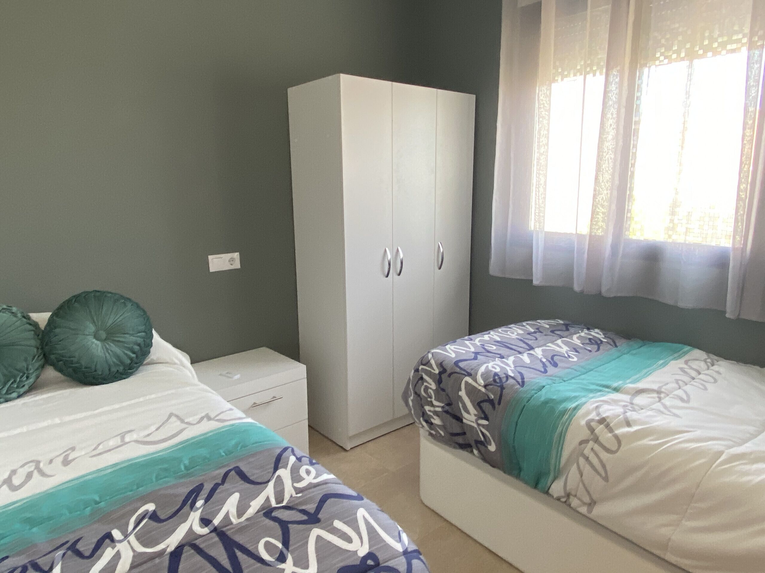 A bedroom with two beds, a wardrobe, and a window with sheer curtains.